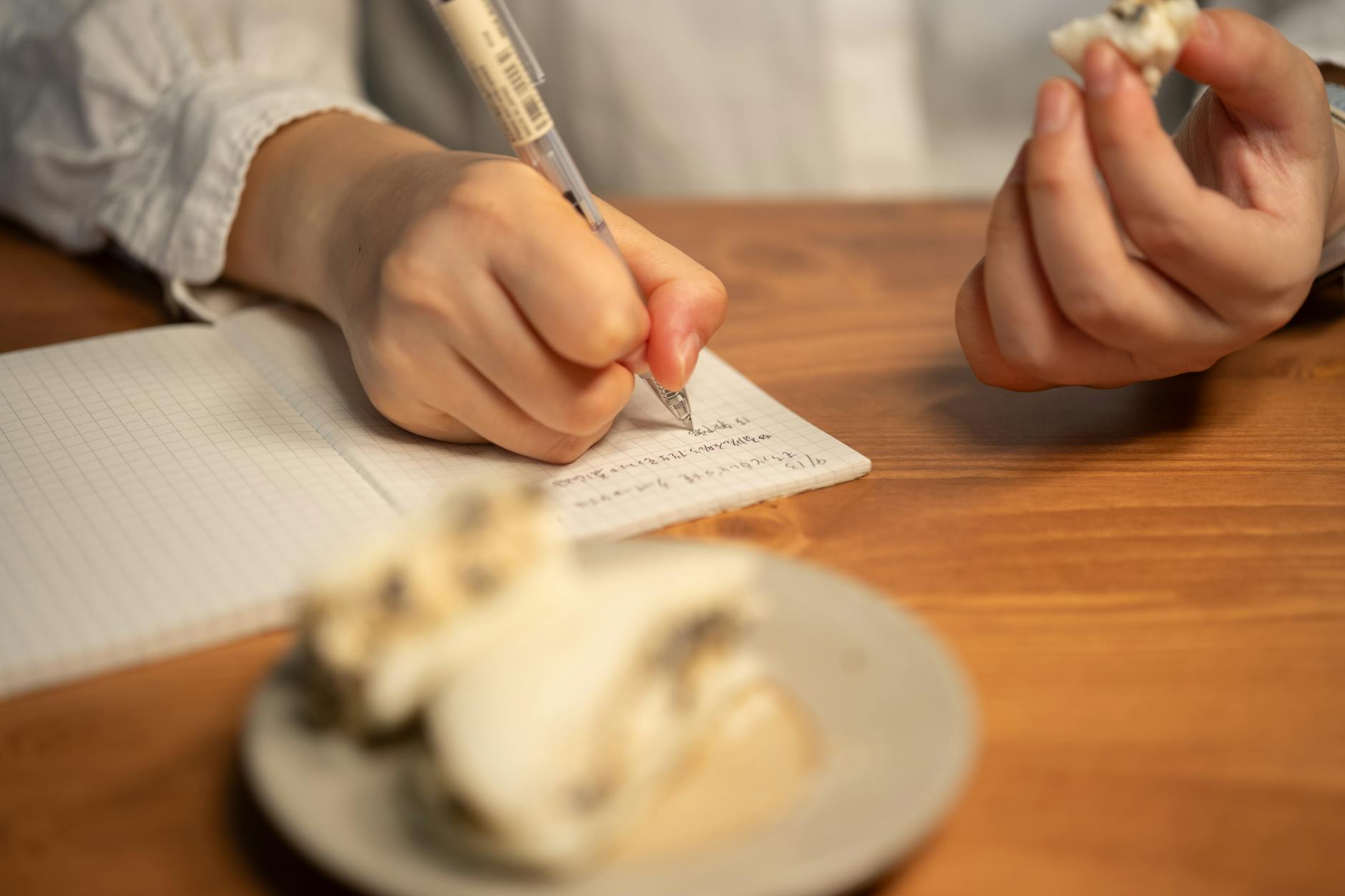 Person writing notes while enjoying a dessert on a wooden table in a cozy setting.